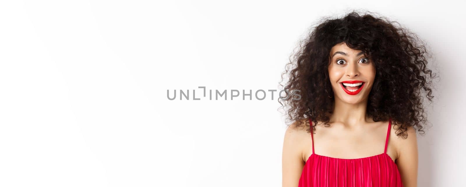 Close-up of happy lady with curly hair and red lips, raising eyebrows and looking surprised at camera, standing over white background.