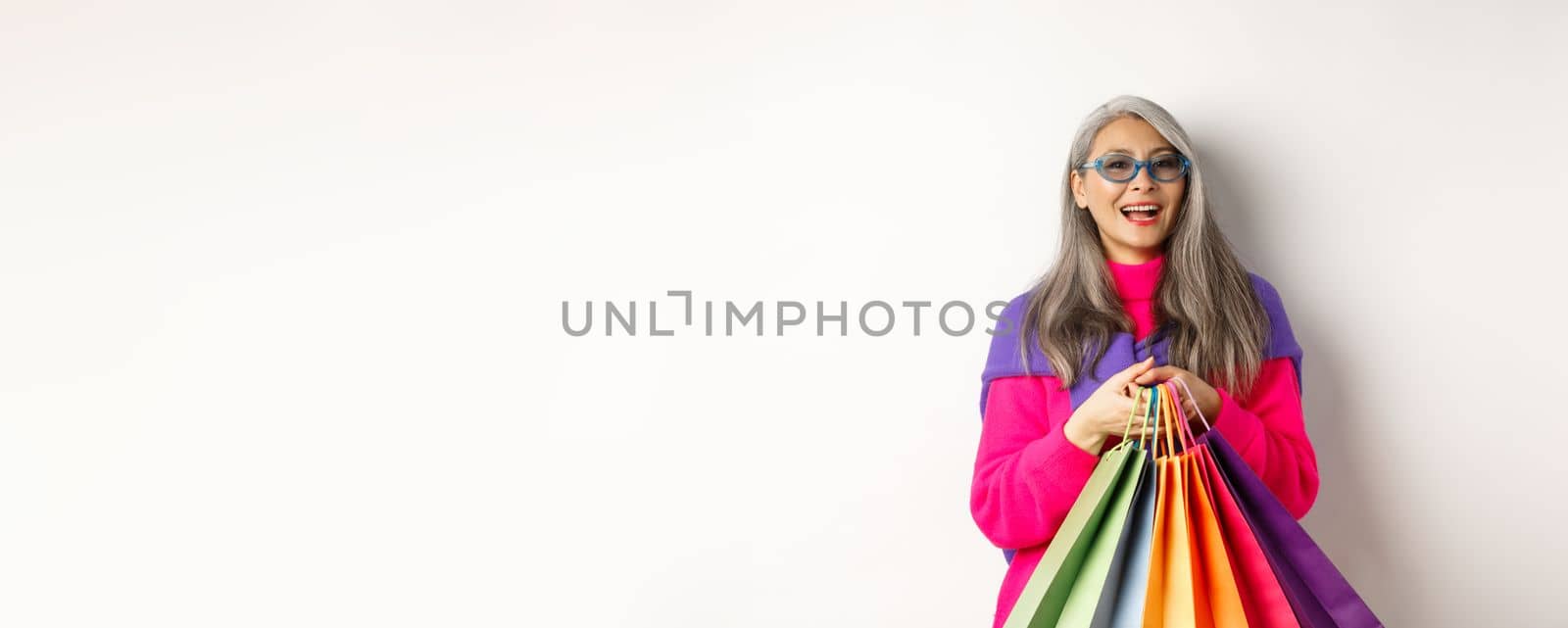 Stylish senior asian woman in sunglasses going shopping on holiday sale, holding paper bags and smiling, standing over white background.