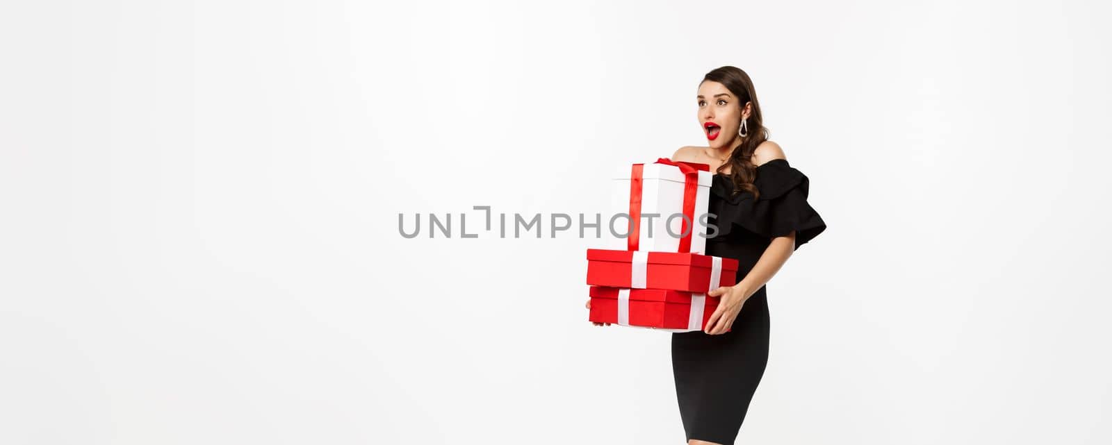 Merry christmas and new year holidays concept. Excited and happy woman in black dress holding xmas presents, looking surprised at logo. standing with presents against white background.