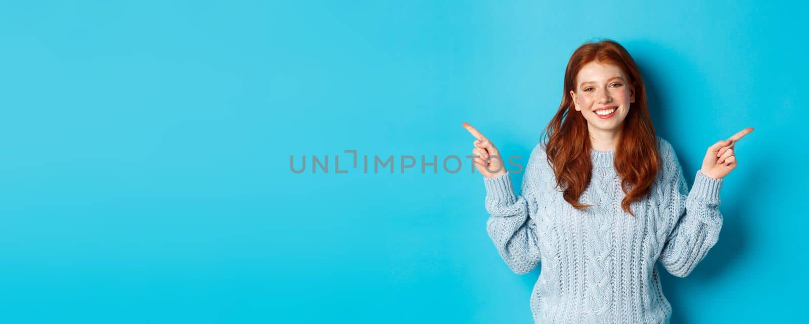 Winter holidays and people concept. Cute teen girl with red hair, smiling and pointing fingers sideways, showing advertisements, standing over blue background by Benzoix