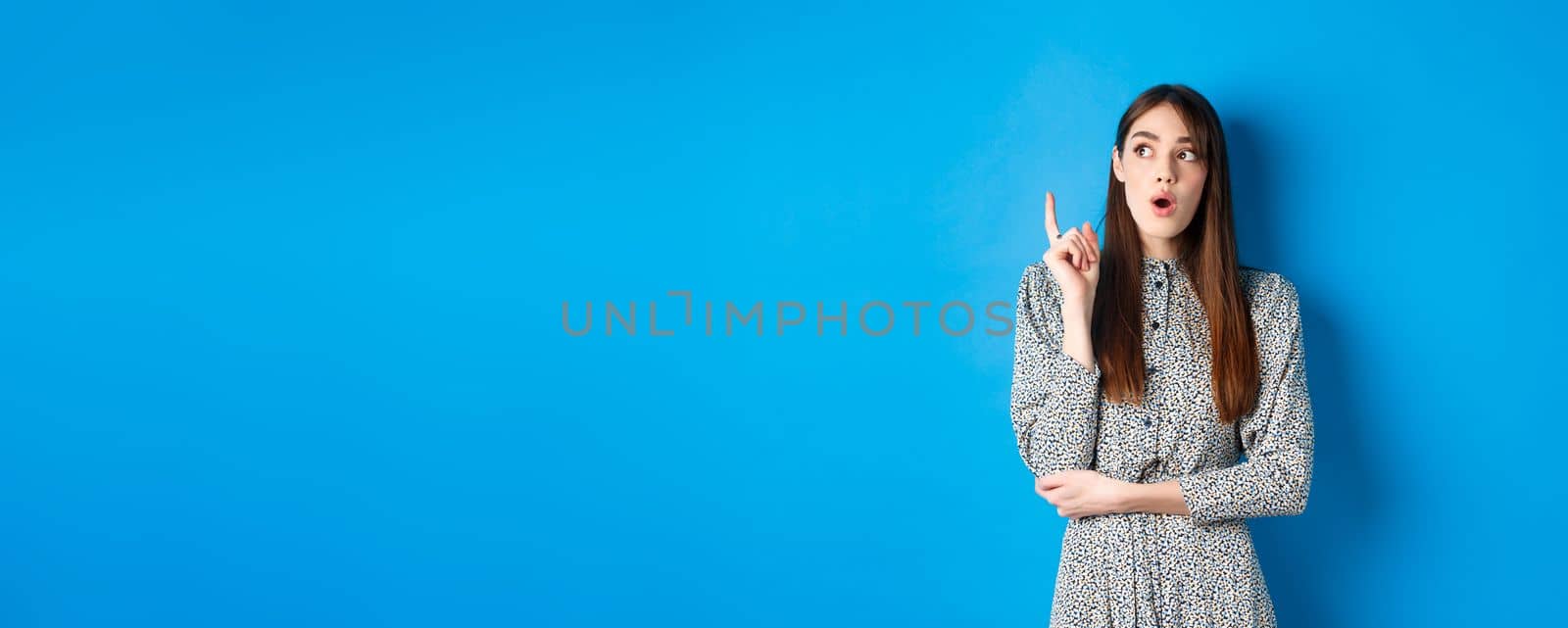 Thoughtful attractive girl in dress pitching an idea, raising finger and looking up, have a plan, standing on blue background by Benzoix