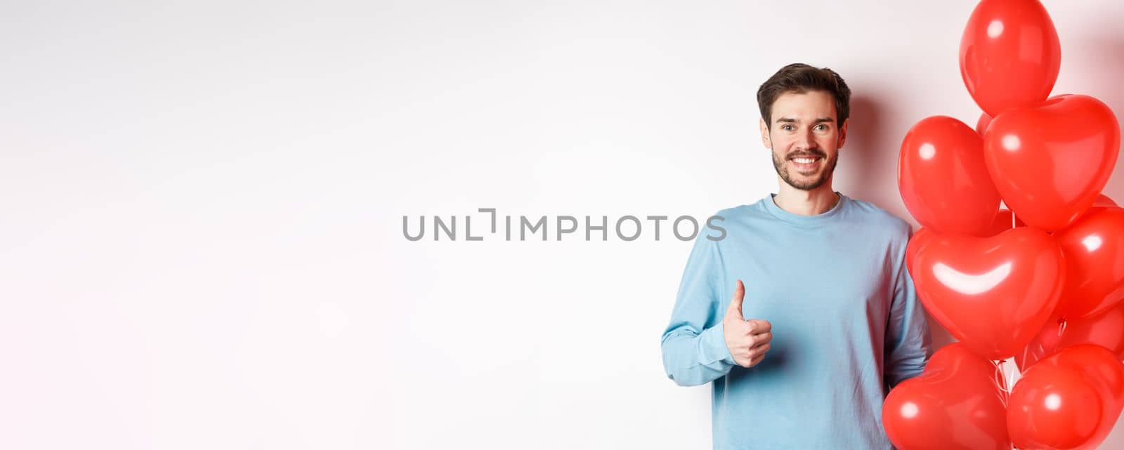 Valentines day and lovers concept. Happy young man showing thumbs up as standing with red hearts balloon, bring romantic gift on date, standing over white background.