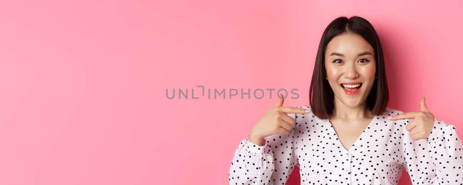 Close-up of happy and confident asian woman pointing at herself, smiling self-assured, standing over pink background by Benzoix