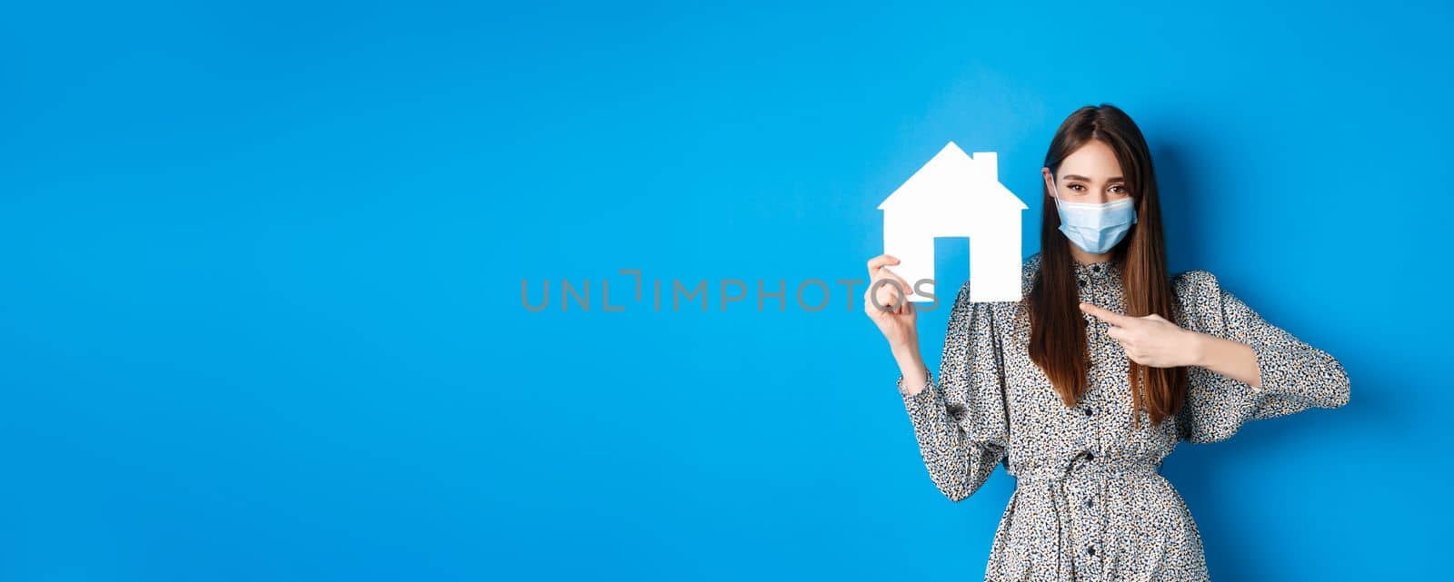 Real estate, covid-19 and pandemic concept. Smiling female model in medical mask pointing at paper house cutout, standing on blue background by Benzoix