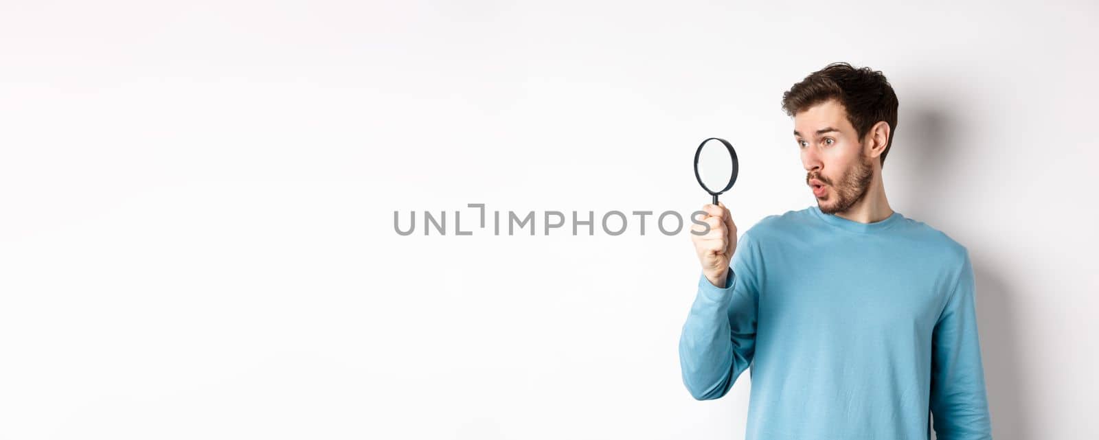 Man got surprised while looking through magnifying glass, saying wow at awesome product, standing over white background by Benzoix