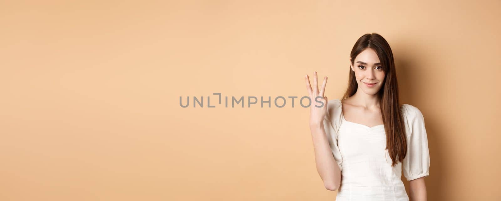 Attractive young woman show fingers number three, smiling and looking confident, standing on beige background.