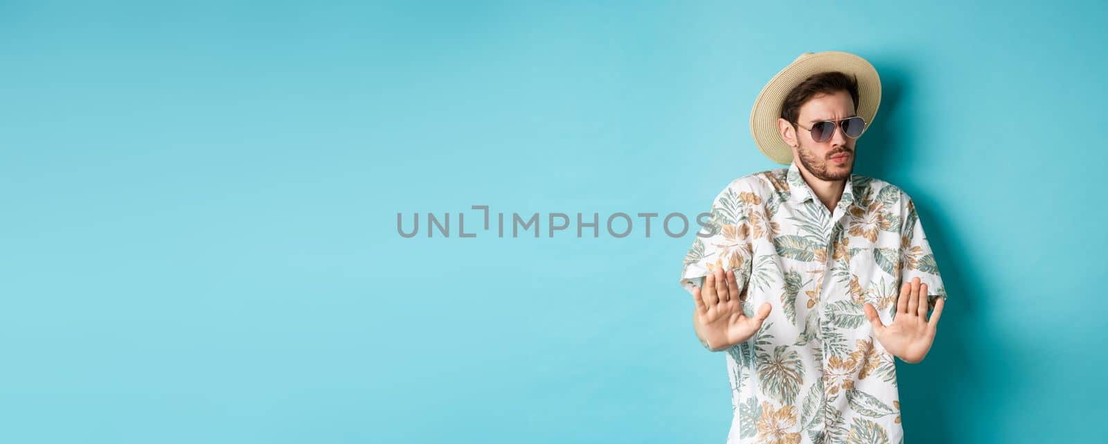 Alarmed tourist asking to stay away, step back from something cringe, showing rejection gesture, standing in straw hat and hawaiian shirt, blue background by Benzoix