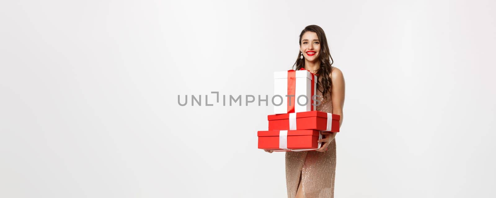 Christmas party and celebration concept. Full-length of stylish woman with red lips, glamour dress, holding gifts and smiling happy, standing over white background by Benzoix