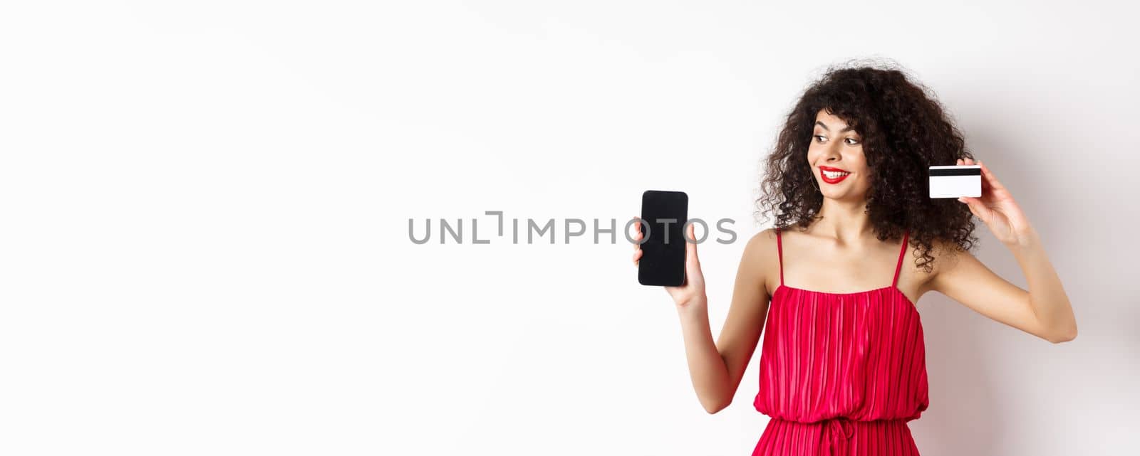 Online shopping concept. Elegant curly-haired woman in red dress showing plastic credit card and empty mobile phone screen, standing over white background.
