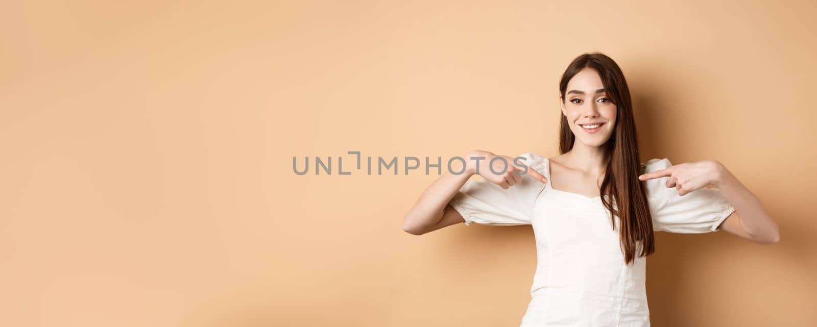Smiling woman pointing at center, showing logo with happy face, standing in white dress on beige background.