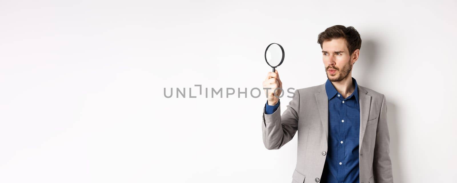 Serious man in suit searching for clues through magnifying glass, investigating, standing on white background.