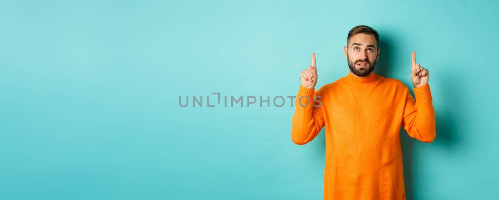 Doubtful and unamused man looking, pointing fingers up something unpleasant, standing skeptical against turquoise background.