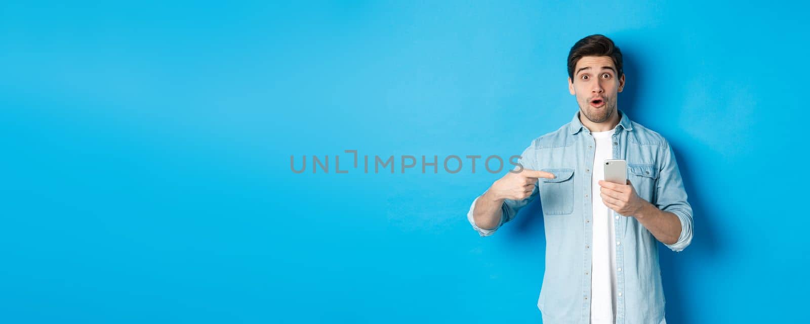 Concept of online shopping, applications and technology. Impressed man pointing finger at mobile phone and looking amazed, recommending app, standing over blue background by Benzoix