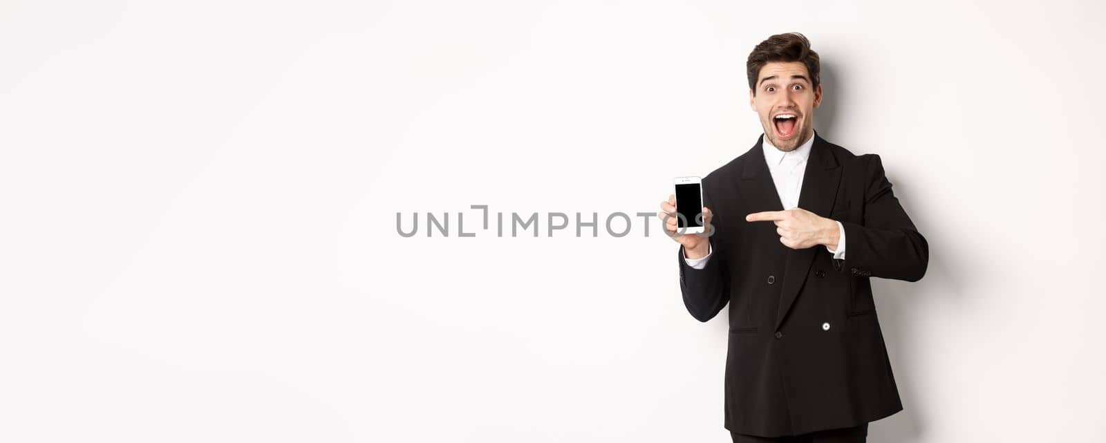 Portrait of handsome businessman in suit, pointing finger at mobile phone screen, showing advertisement, standing over white background.