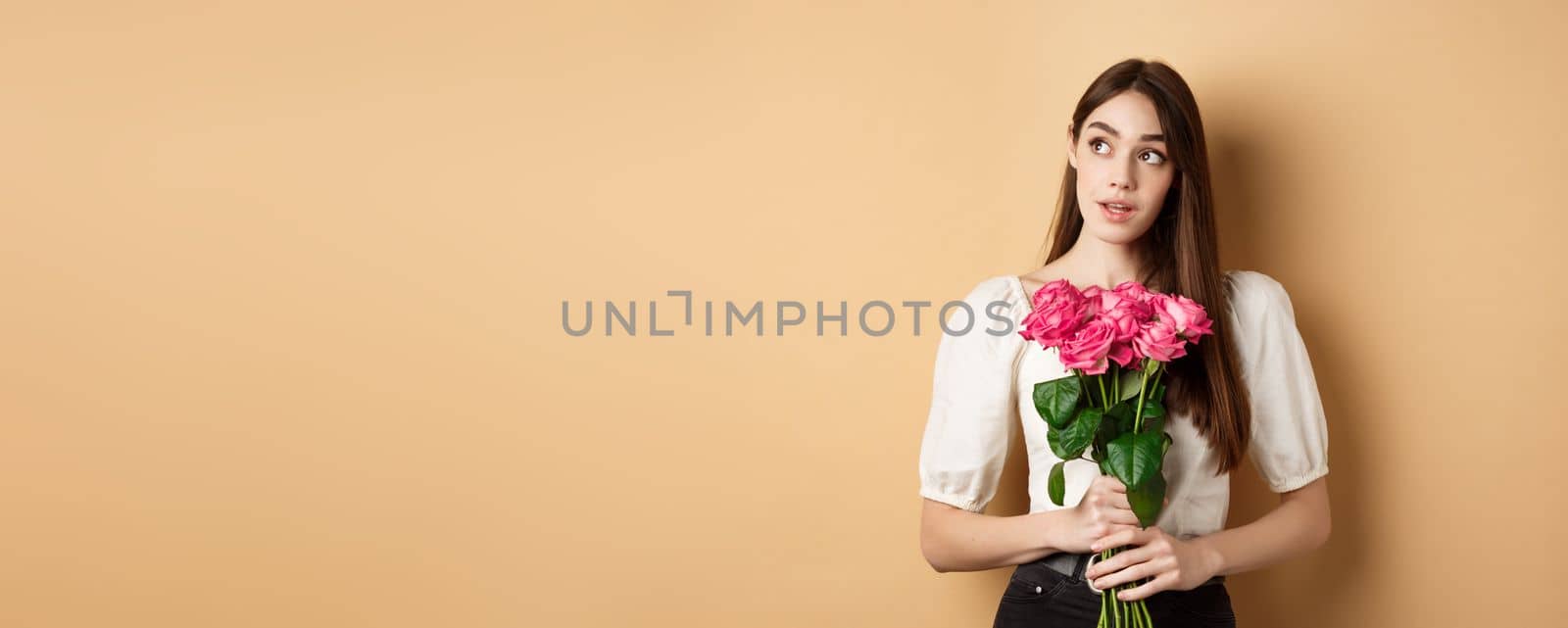 Romantic Valentines day concept. Dreamy woman with bouquet of pink rose, looking aside at logo, holding flowers on beige background.