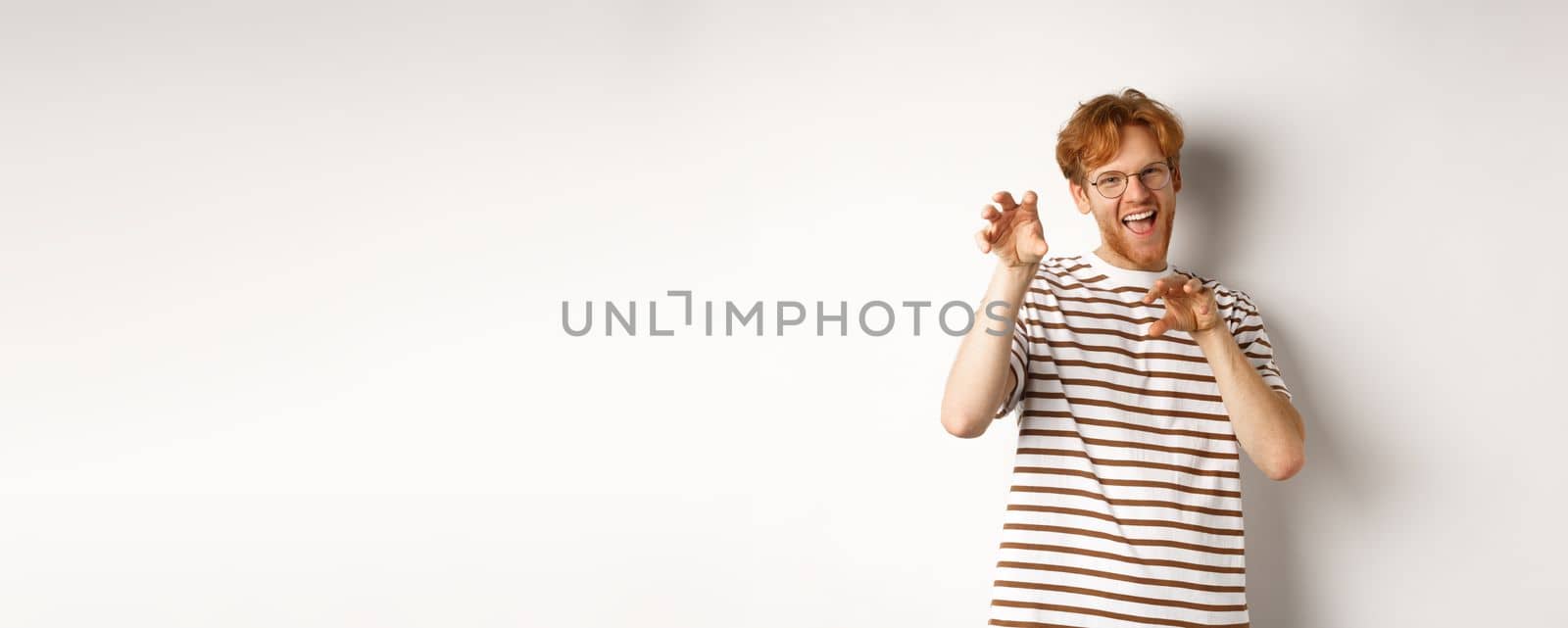 Funny young man with red hair and beard showing claws gesture and roaring, having fun, standing happy over white background.