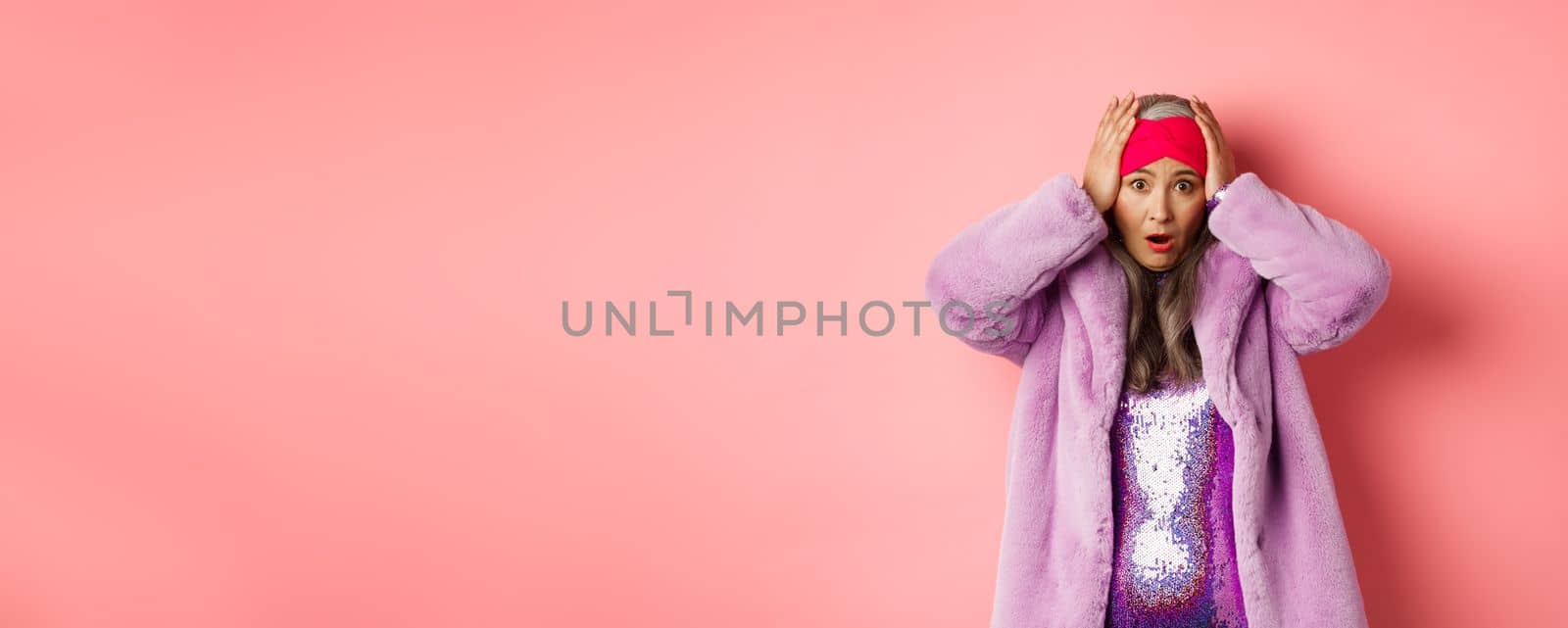 Shocked and alarmed asian senior woman, wearing purple faux fur coat, holding hands on head and panicking, standing distressed against pink background by Benzoix