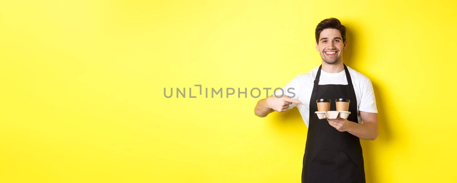 Handsome barista holding two cups of takeaway coffee, pointing finger at drinks and smiling, standing in black apron against yellow background by Benzoix