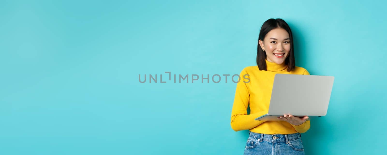 Happy asian woman in yellow pullover using laptop, shopping online or working, standing over blue background.