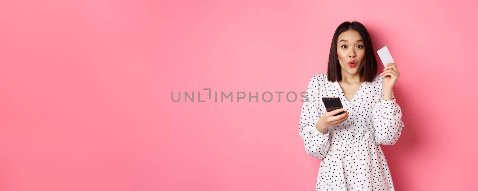 Attractive young asian woman order online, holding credit card and mobile phone, making internet purchase, standing happy over pink background.