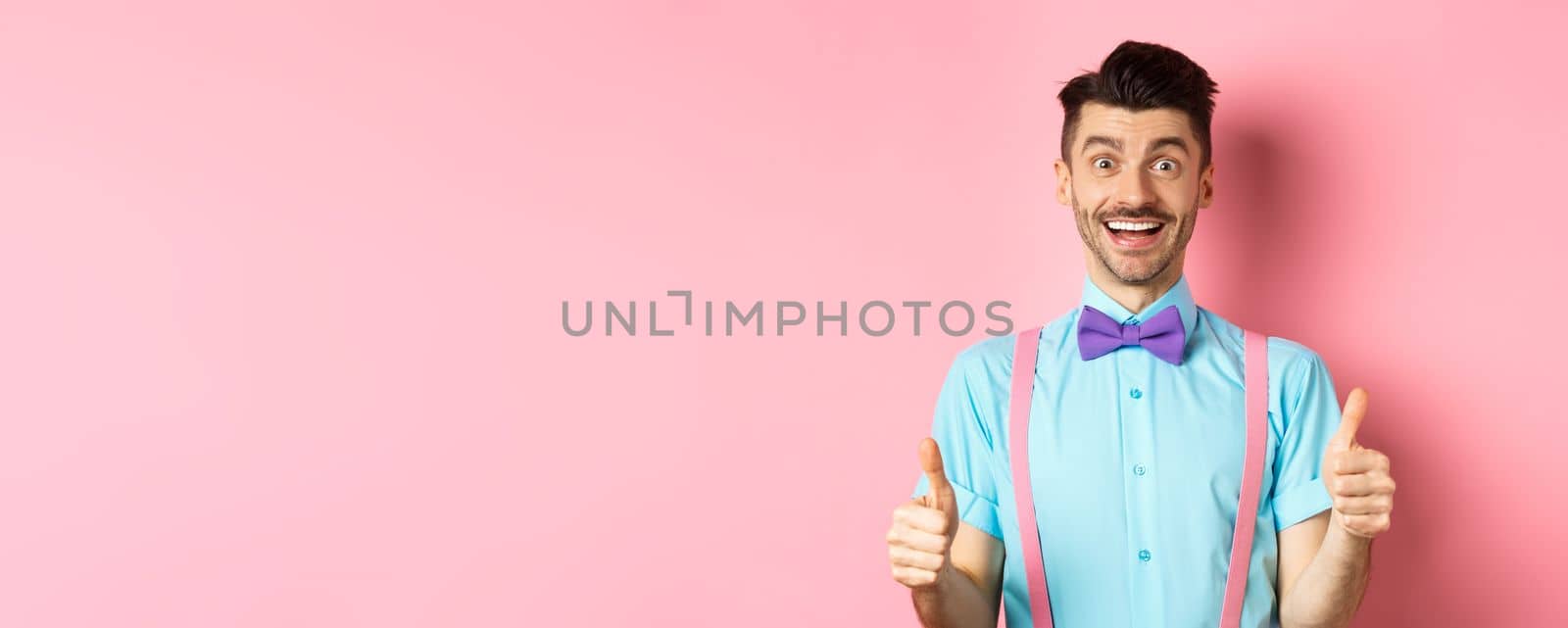 Excited caucasian guy showing thumbs up and praising great work, well done gesture, standing in suspenders and bow-tie on pink background by Benzoix