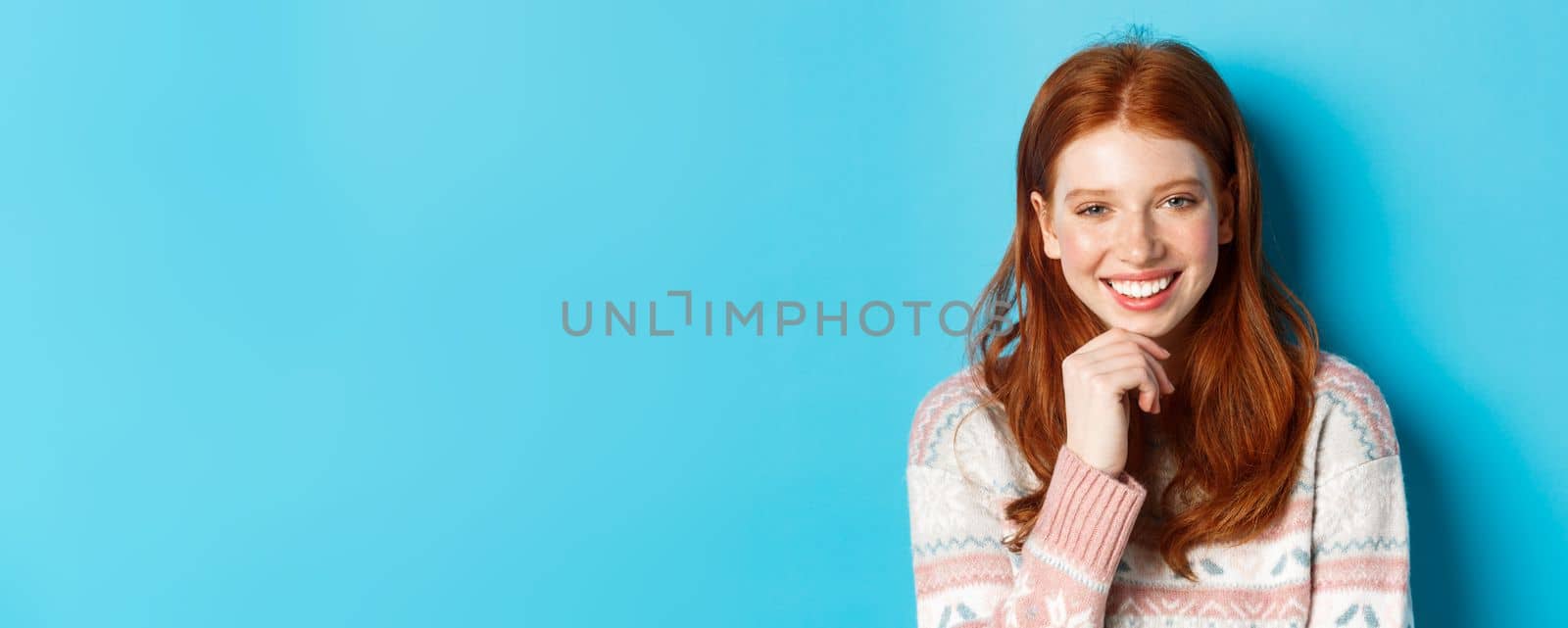 Close-up of beautiful redhead girl smiling, having conversation and staring at camera interested, standing over blue background by Benzoix