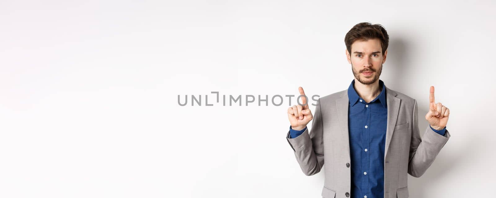 Serious businessman with beard wearing suit, pointing fingers up, look here gesture, advertising on white background.