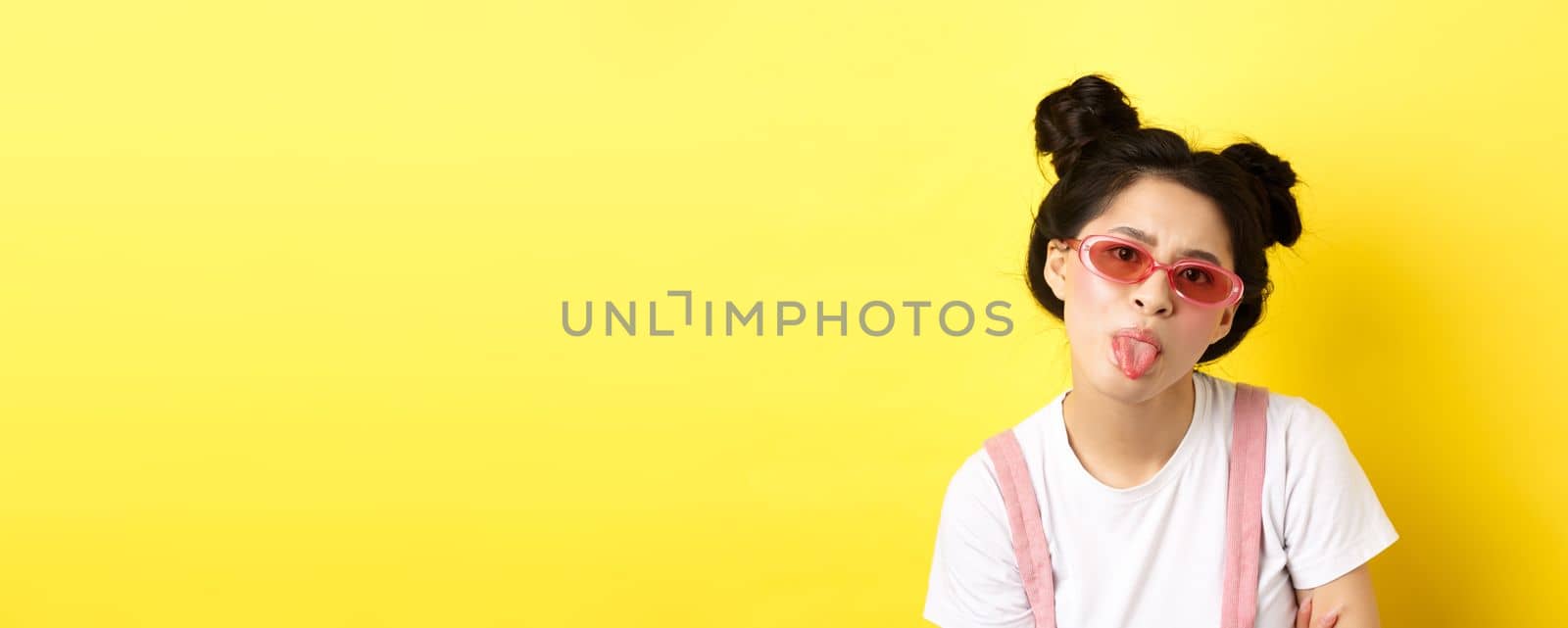 Summer and beauty concept. Childish asian girl in sunglasses showing tongue, being rude, standing on yellow background by Benzoix