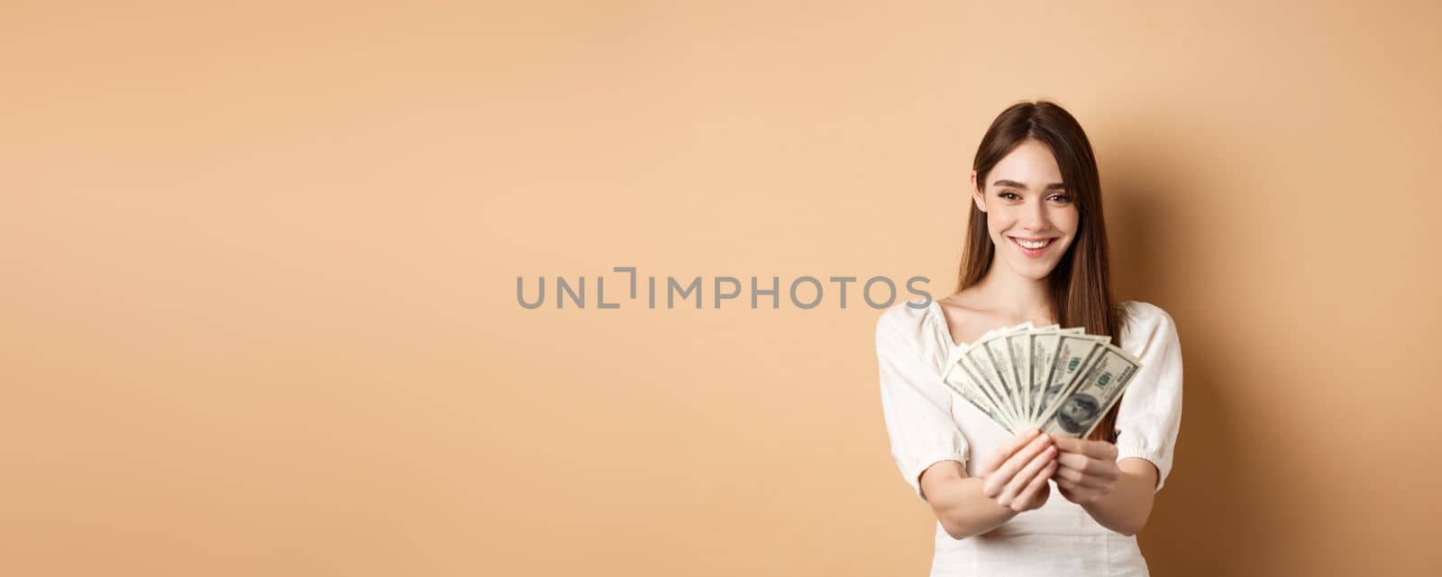 Young woman showing dollar bills and smiling, standing with money on beige background. Concept of loan and insurance.