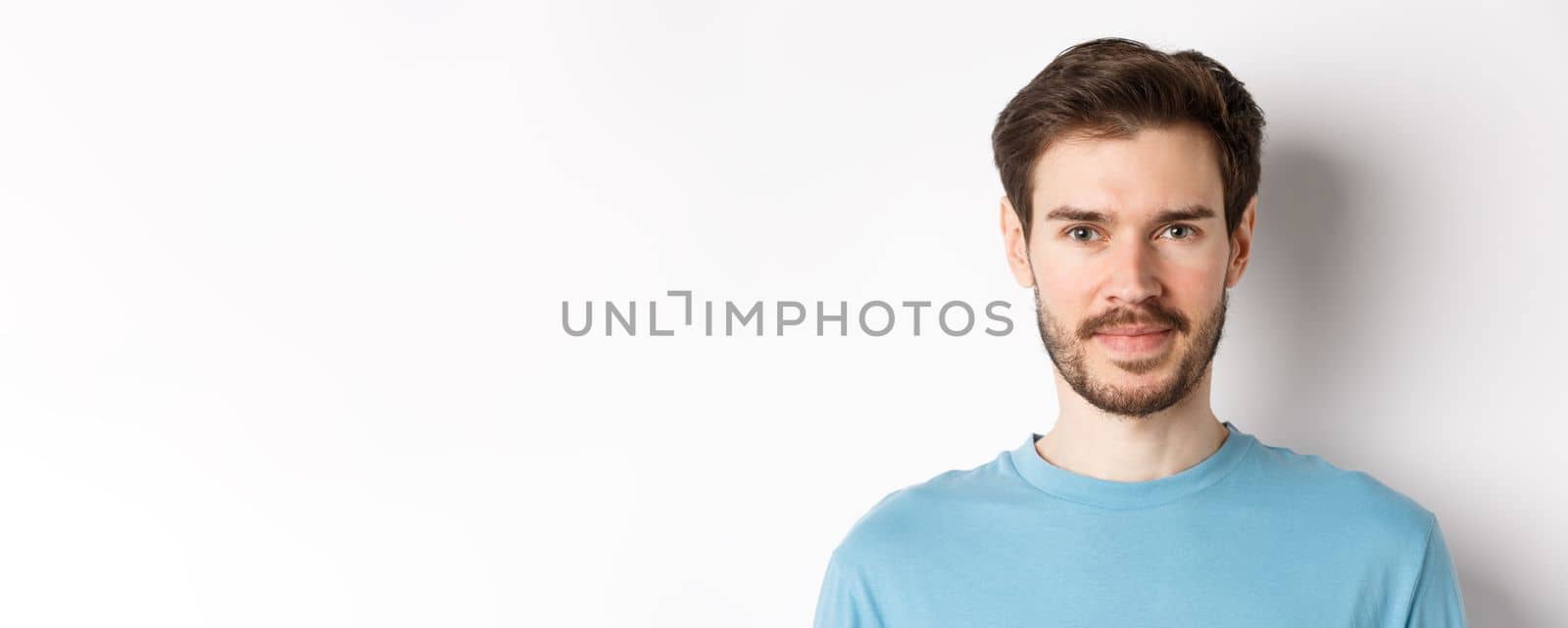 Close up of young handsome man with beard, smiling at camera with confidence, standing over white background by Benzoix