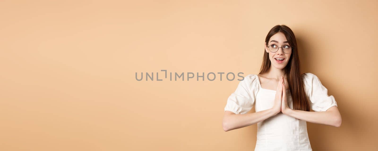 Hopeful girl holding hands in pray, begging for dream come true, looking aside excited, standing on beige background. Copy space