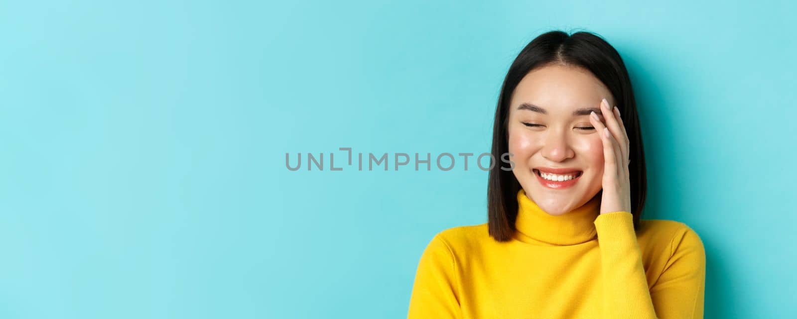 Beauty and makeup concept. Close up of beautiful asian woman laughing and looking flushed from compliments, standing over blue background.