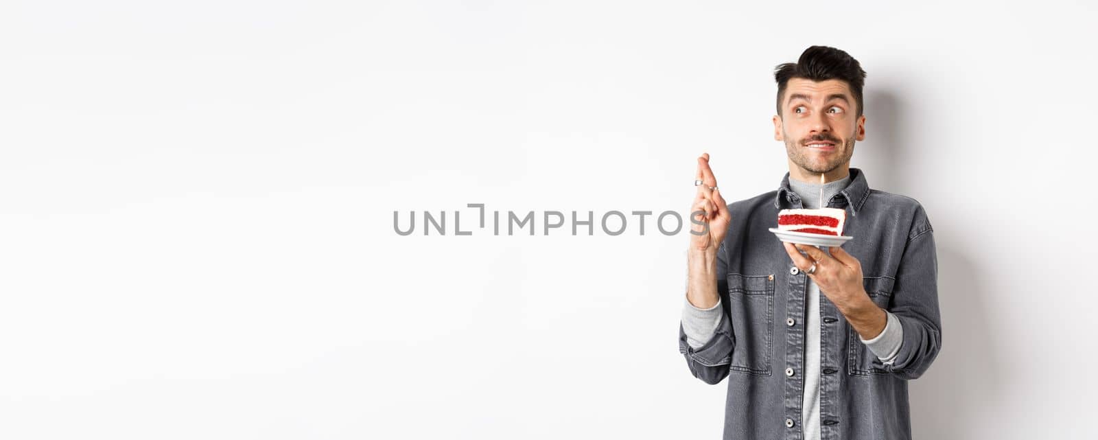 Birthday. Happy young man celebrating, making wish with cake and cross fingers, looking hopeful aside, standing on white background by Benzoix