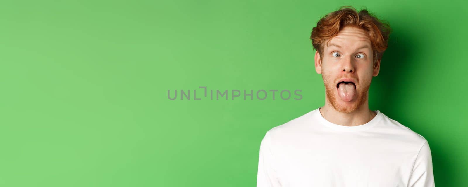 Emotions and fashion concept. Close up of funny redhead man showing silly faces, sticking tongue and squinting, standing over green background.