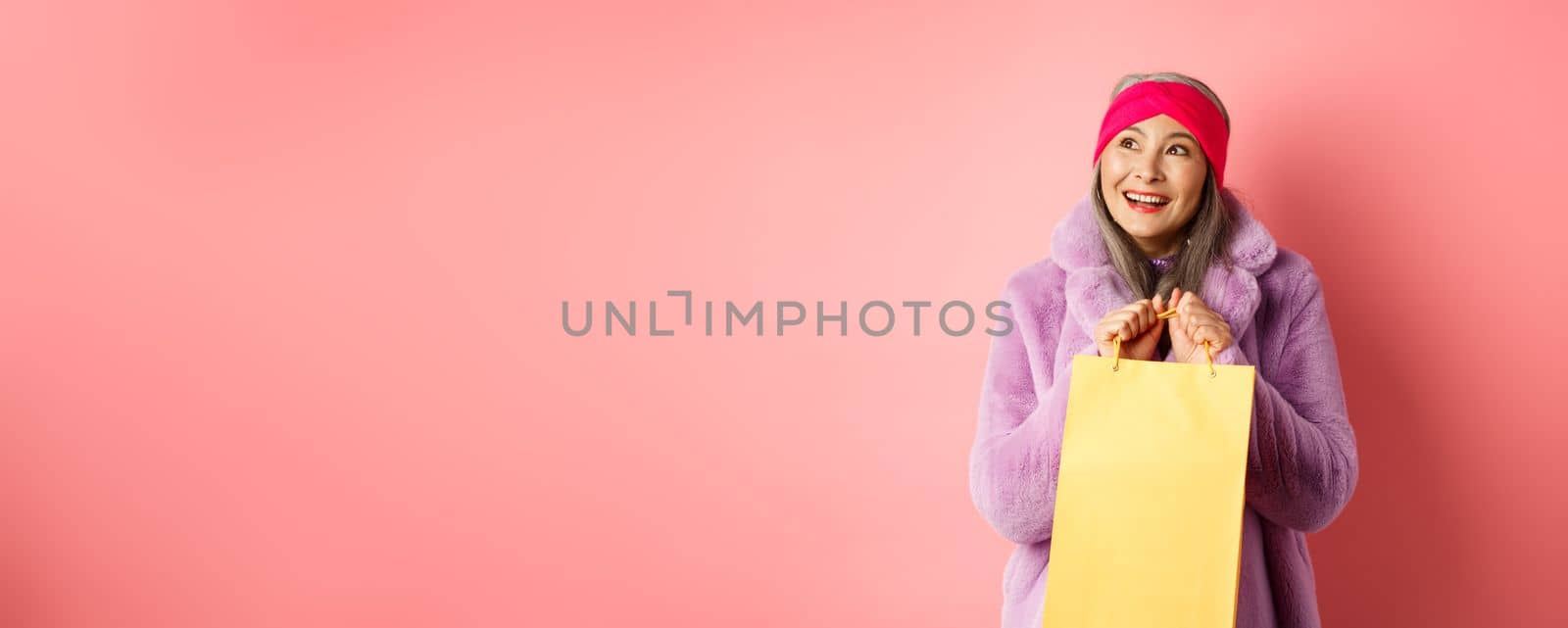 Shopping and fashion concept. Beautiful asian senior woman looking dreamy at upper left corner and imaging something, holding yellow paper bag with gift, pink background.