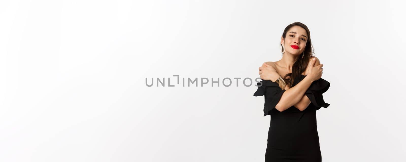 Fashion and beauty. Sensual and tender woman in black dress, embracing own body, hugging herself and smiling, standing over white background.