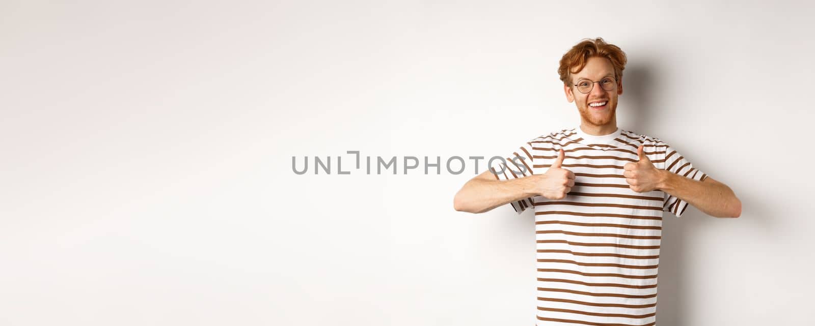 Cheerful redhead guy in nerdy glasses showing thumbs-up, smiling and saying yes, agree or like something, standing over white background.