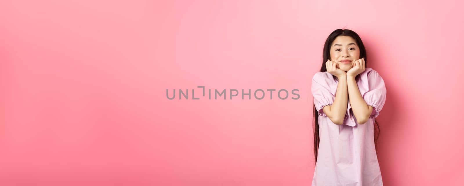 Cute and happy asian woman waiting for something with excitement, lean face on hands and watching lovely scene with admiration, standing against pink background.