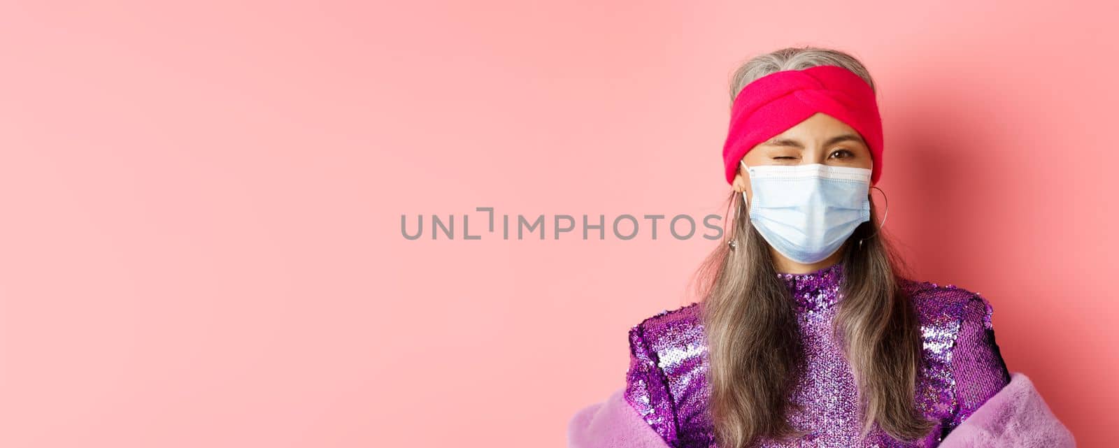 Covid-19, virus and social distancing concept. Sassy asian senior woman in medical mask and glitter dress winking, wearing party outfit, pink background by Benzoix