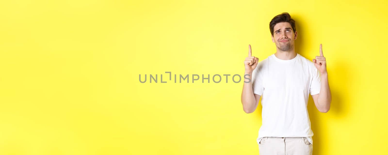 Skeptical young man pointing and looking up at something bad, judging offer, standing over yellow background.