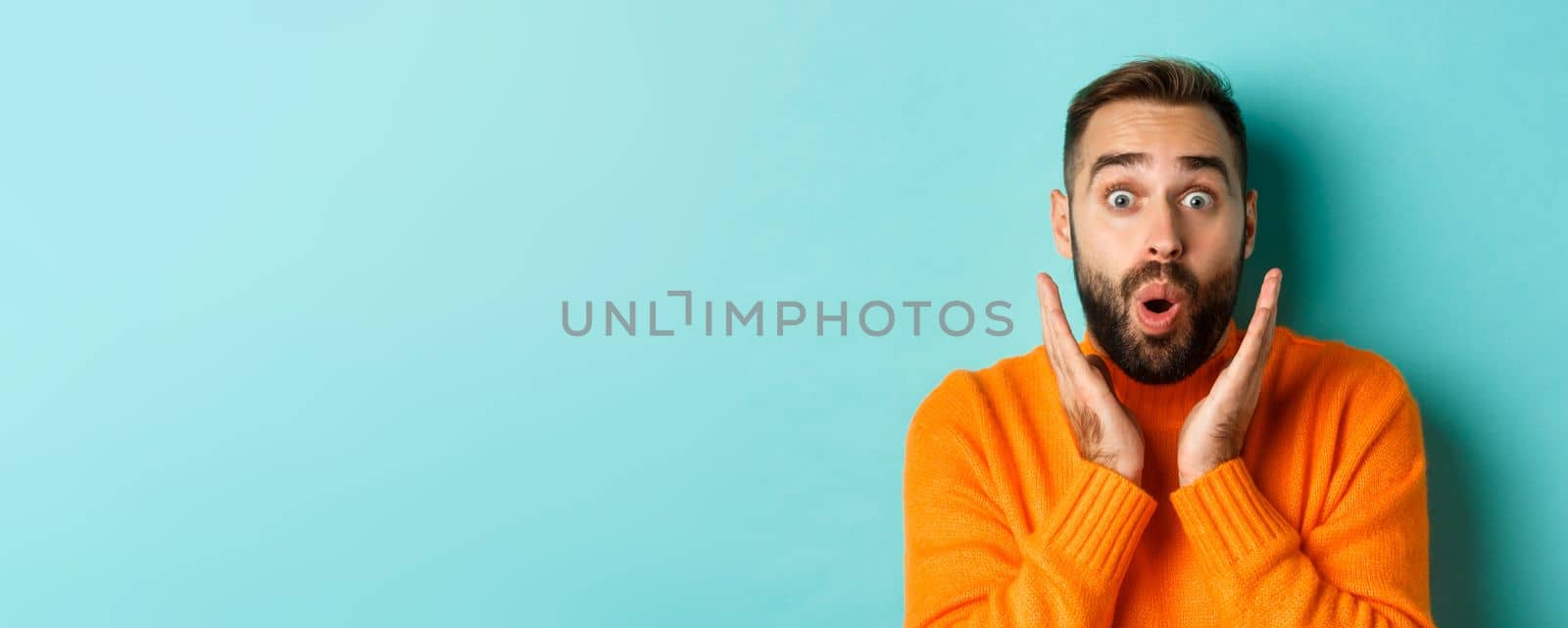 Close-up of handsome caucasian man staring at camera surprised, wearing orange sweater, standing against turquoise background.