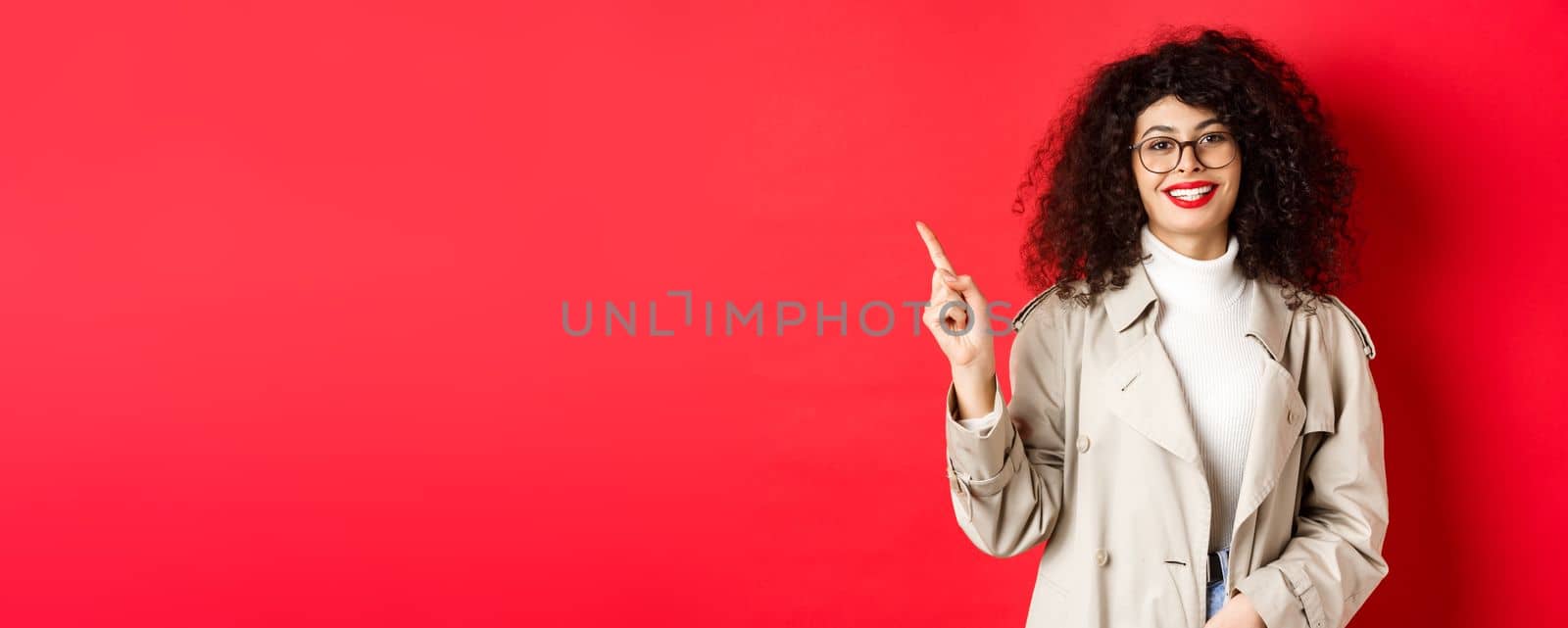 Portrait of stylish caucasian woman in trench coat and glasses pointing finger aside, showing empty space on red background and smiling by Benzoix