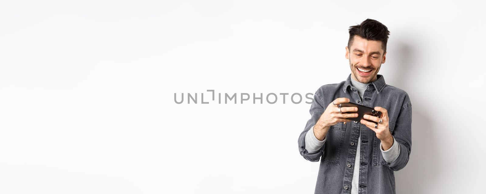 Handsome young man playing mobile video game and smiling at screen, standing against white background by Benzoix