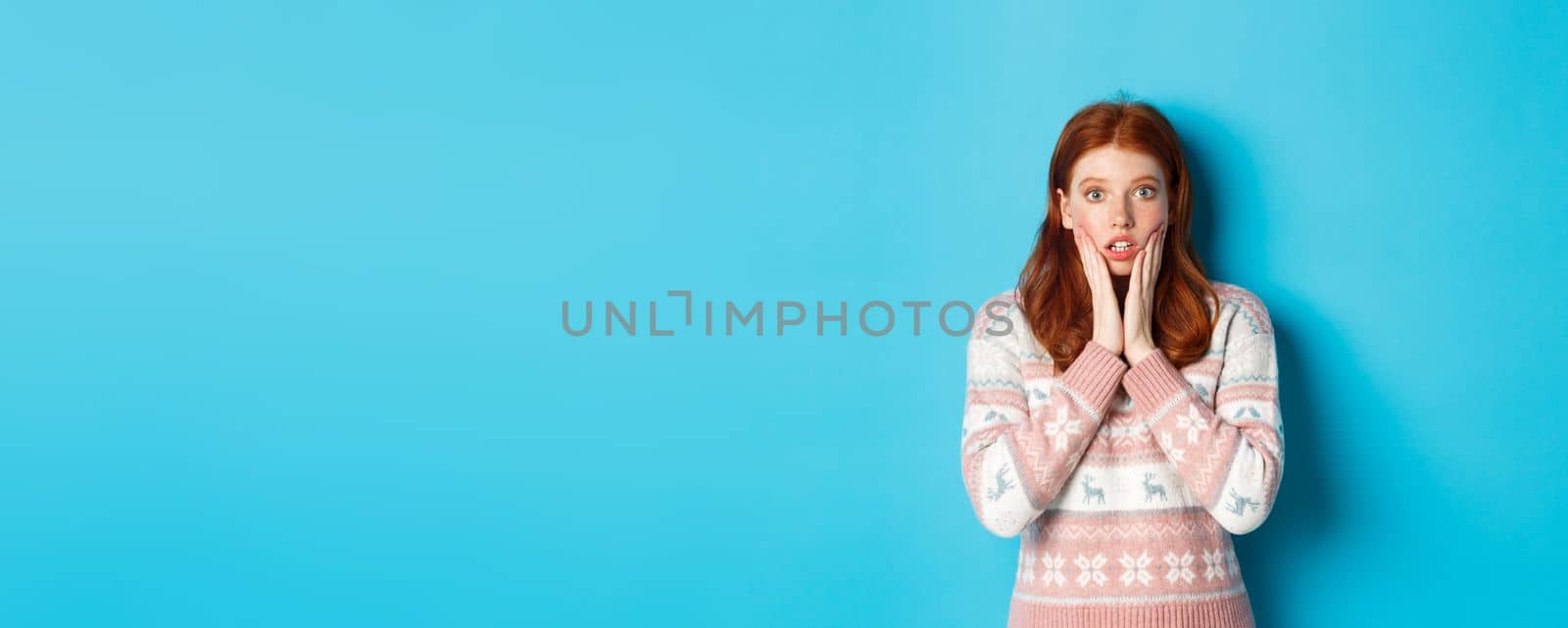 Image of impressed redhead girl gasping and staring at camera, hear big news, standing in winter sweater against blue background.