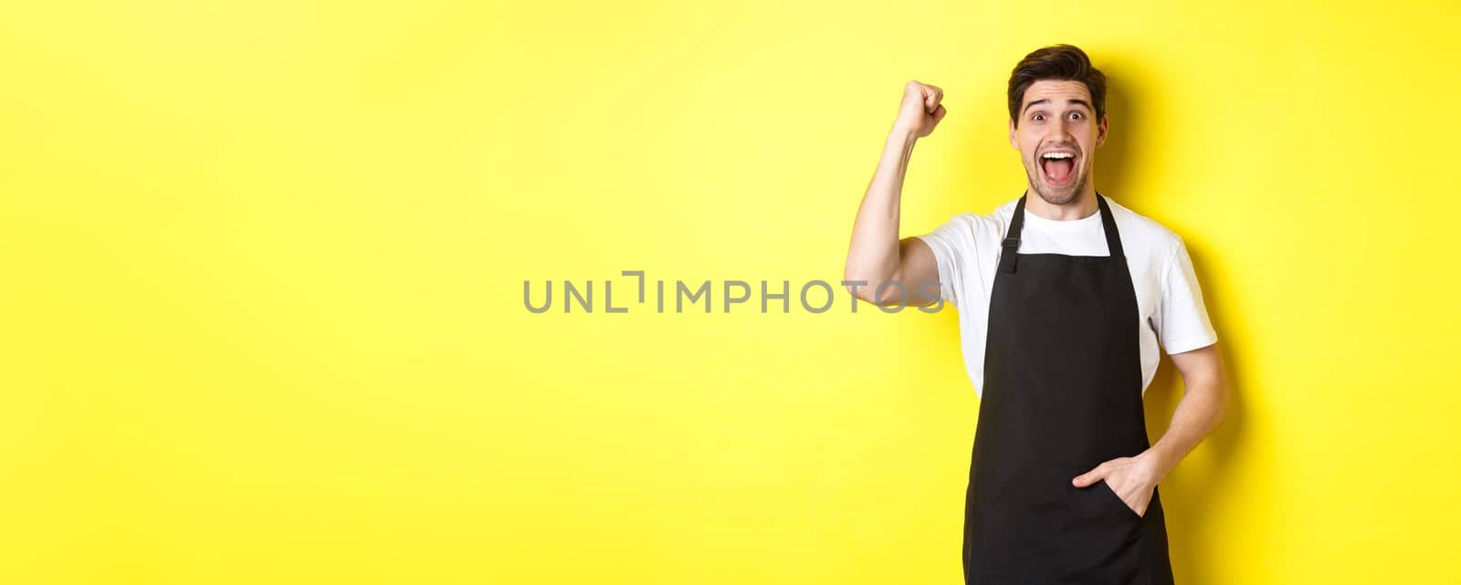 Cheerful seller making fist pump, rejoicing and triumphing, standing in black apron against yellow background.