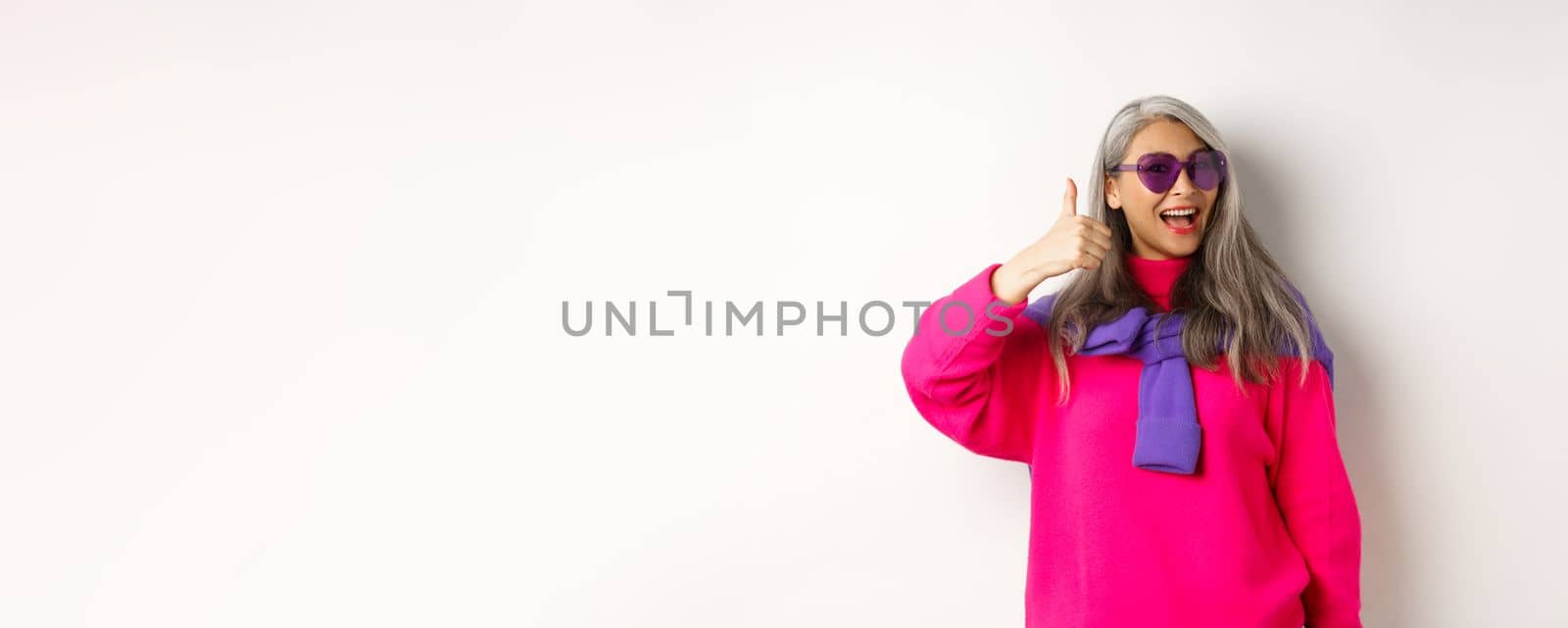 Shopping concept. Stylish asian senior woman in sunglasses and trendy outfit, showing thumb-up in approval, recommending shop, standing over white background by Benzoix