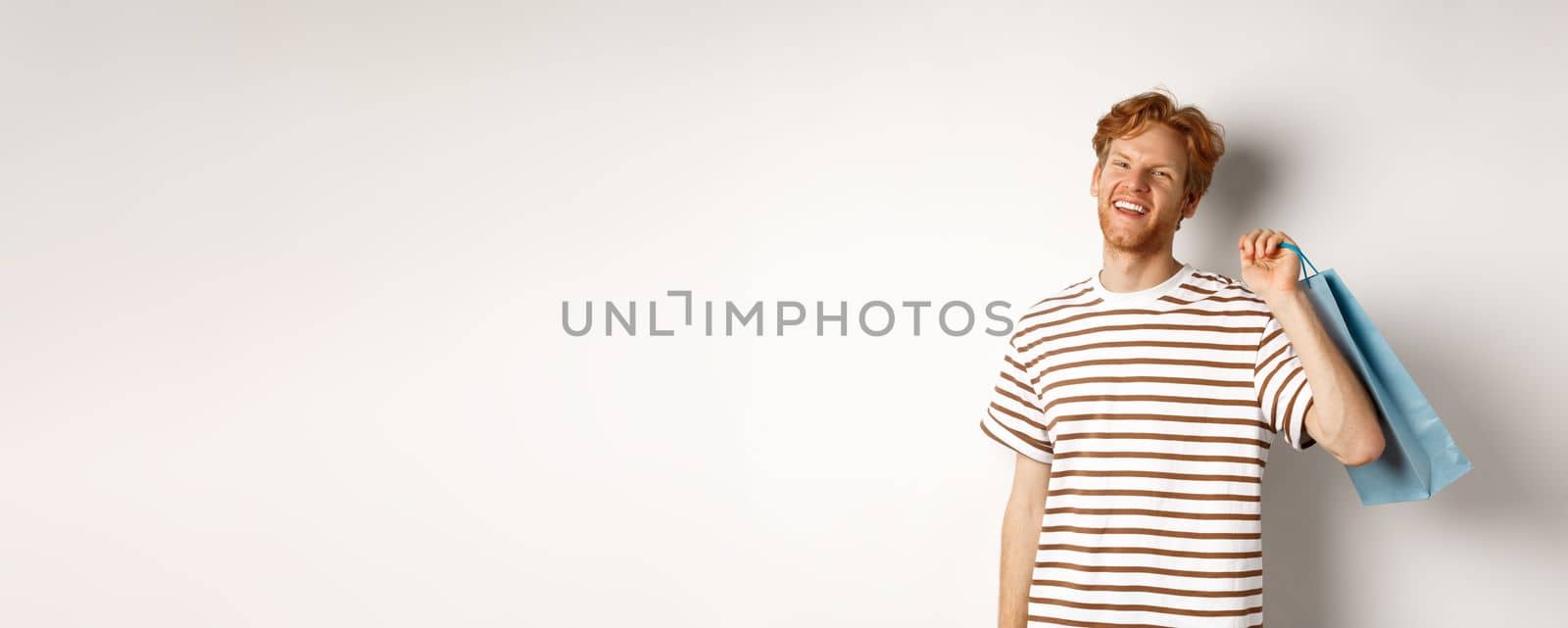 Cheerful bearded man with red hair enjoying shopping, holding bag over shoulder and smiling, standing over white background by Benzoix