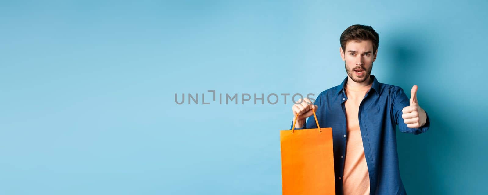 Cool guy showing thumb up and holding orange shopping bag, standing on blue background.