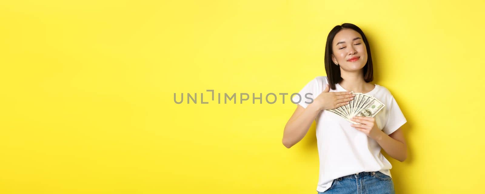 Beautiful asian woman love money, hugging dollars and smiling pleased, standing over yellow background.