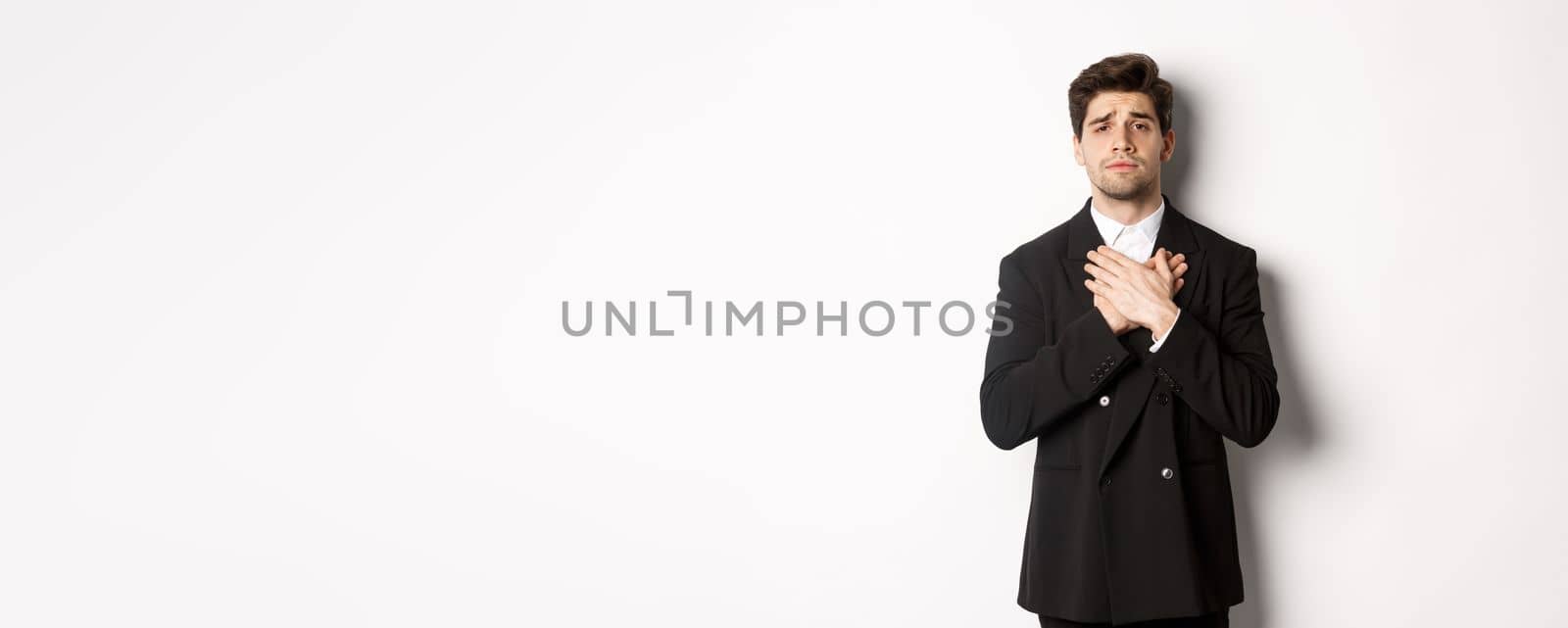 Portrait of touched and compassionate guy in suit, holding hands on heart and looking with pity at camera, standing over white background.